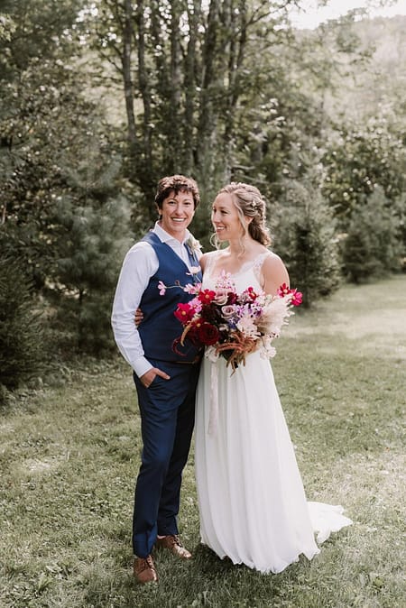 A beautiful same sex couple standing together outside embracing after their ceremony, holding an artful bridal bouquet with dried pampas grass designed by Nectar and Root, Vermont wedding florist.