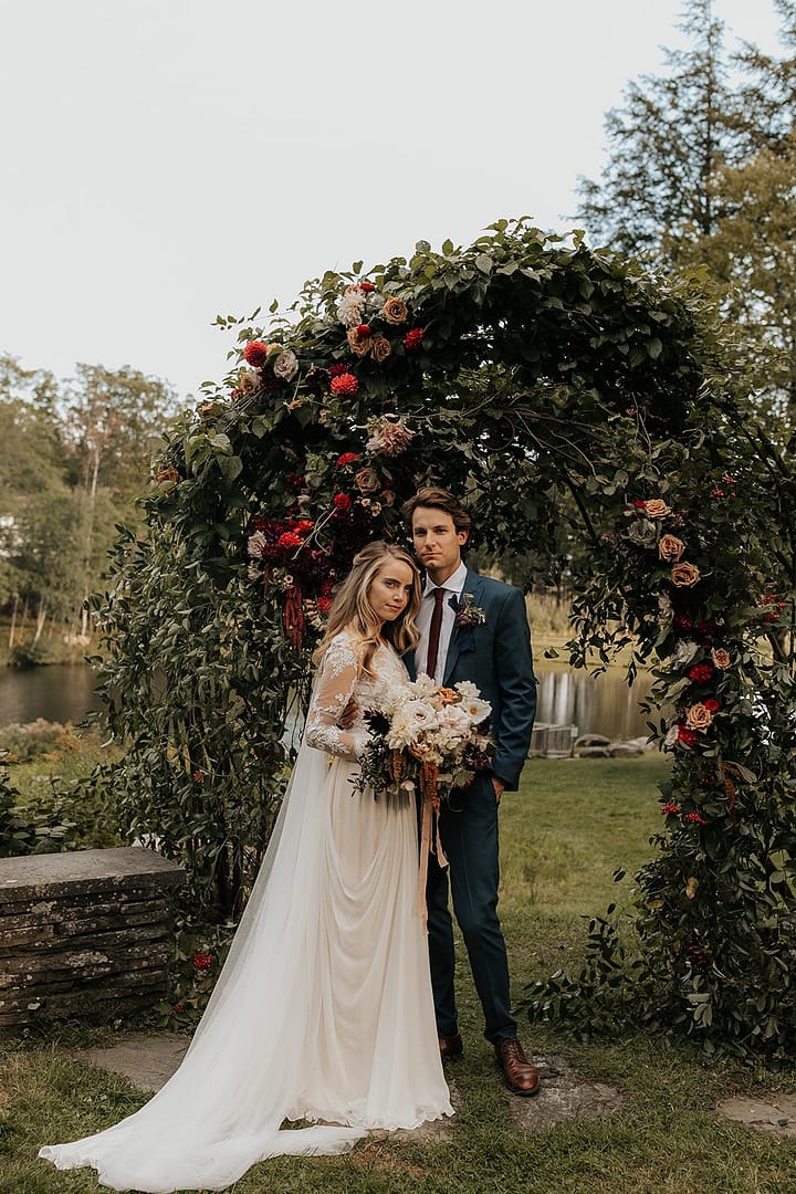 Bride and groom standing beside romantic outdoor fall ceremony arbor while holding a modern September bridal bouquet of garden roses, hanging amaranth, sweet peas, dahlias, cosmos and zinnias by Nectar and Root, Vermont wedding florist at Edson Hill in Stowe, Vermont.
