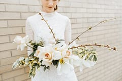 A bride wearing a short mock neck wedding dress holding an artful spring bridal bouquet of May tulips, garden roses, sweet peas, and flowering branches in a warm dusty neutral color palette by Nectar and Root, Vermont wedding florist.
