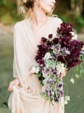 A bride outside wearing a taupe organic wedding dress holding a summer bridal bouquet of July sweet peas in a purple color palette with flowing silk ribbons by Nectar and Root, Vermont wedding florist at Lake Willoughby.