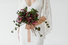 A bride in a rue de seine wedding dress holding a simple modern purple bridal bouquet of spring May hellebores with flowing hand dyed silk ribbon by Nectar and Root, Vermont wedding florist.