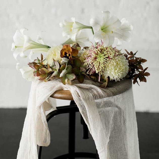 A modern spring reception centerpiece in a warm neutral palette atop an industrial table with cheesecloth tablecloth featuring lilies, pansies, mums and hellebores by Nectar and Root, Vermont wedding florist.