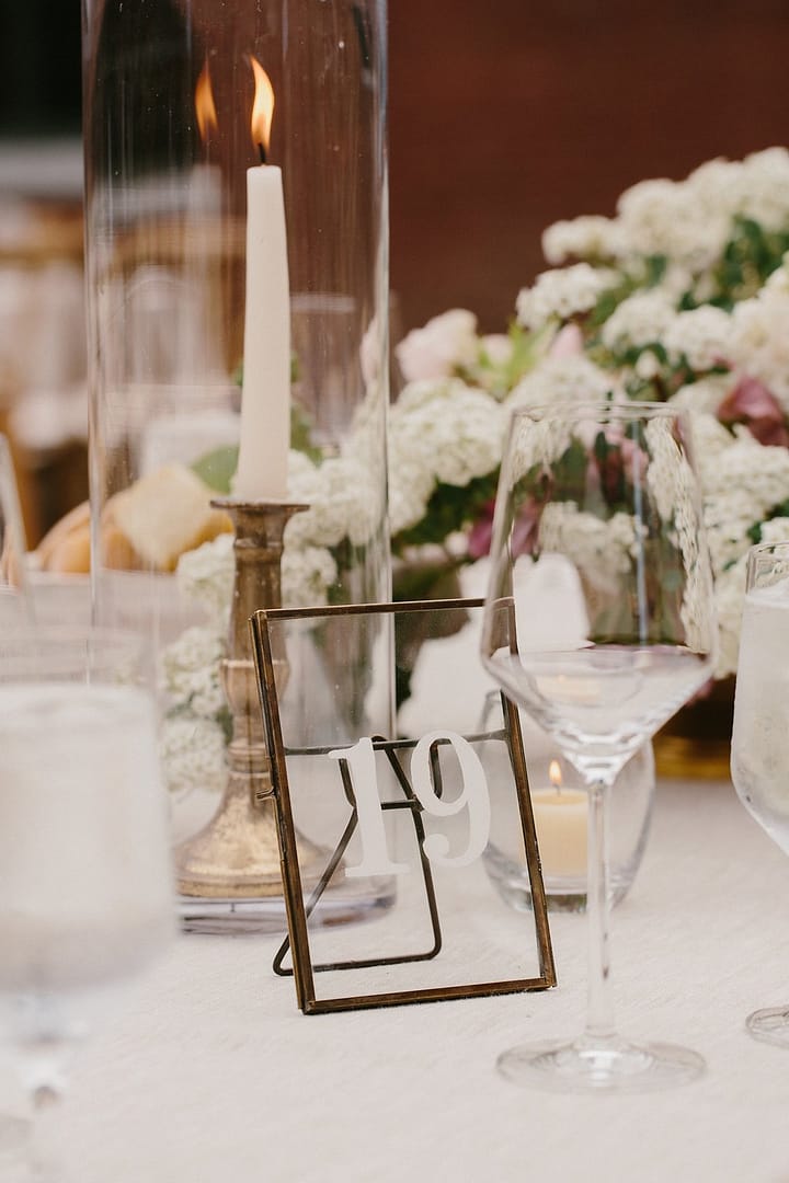 White centerpiece next to tapered candlesticks, gold candleholders and gold table number by Nectar and Root, Vermont wedding florist at Shelburne Coach Barn in Shelburne, Vermont.
