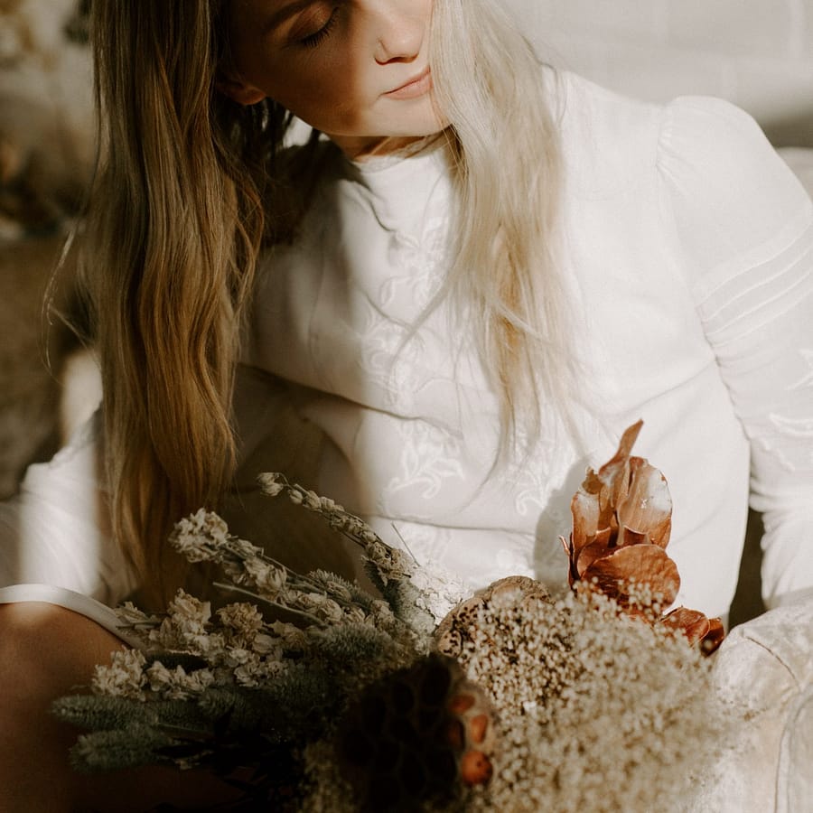 A bride holding a modern bridal fall bridal bouquet of dried flowers in a warm dusty neutral color palette in the sunshine by Nectar and Root, Vermont wedding florist.