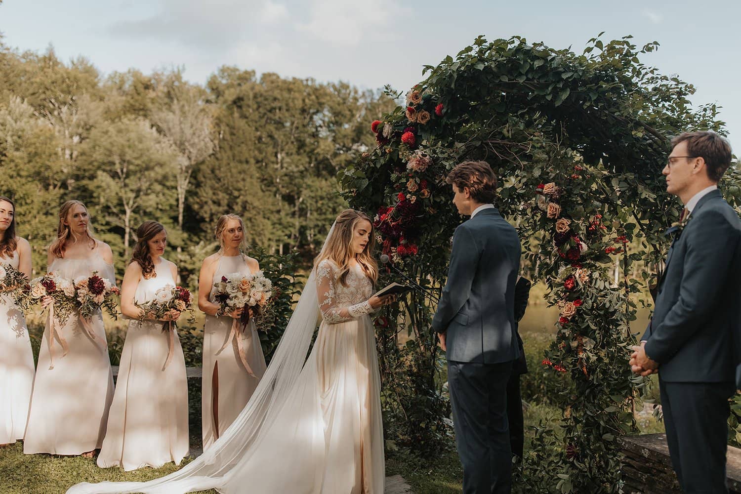 Bride and groom standing beside romantic outdoor fall ceremony arbor while holding a modern September bridal bouquet of garden roses, hanging amaranth, sweet peas, dahlias, cosmos and zinnias by Nectar and Root, Vermont wedding florist at Edson Hill in Stowe, Vermont.