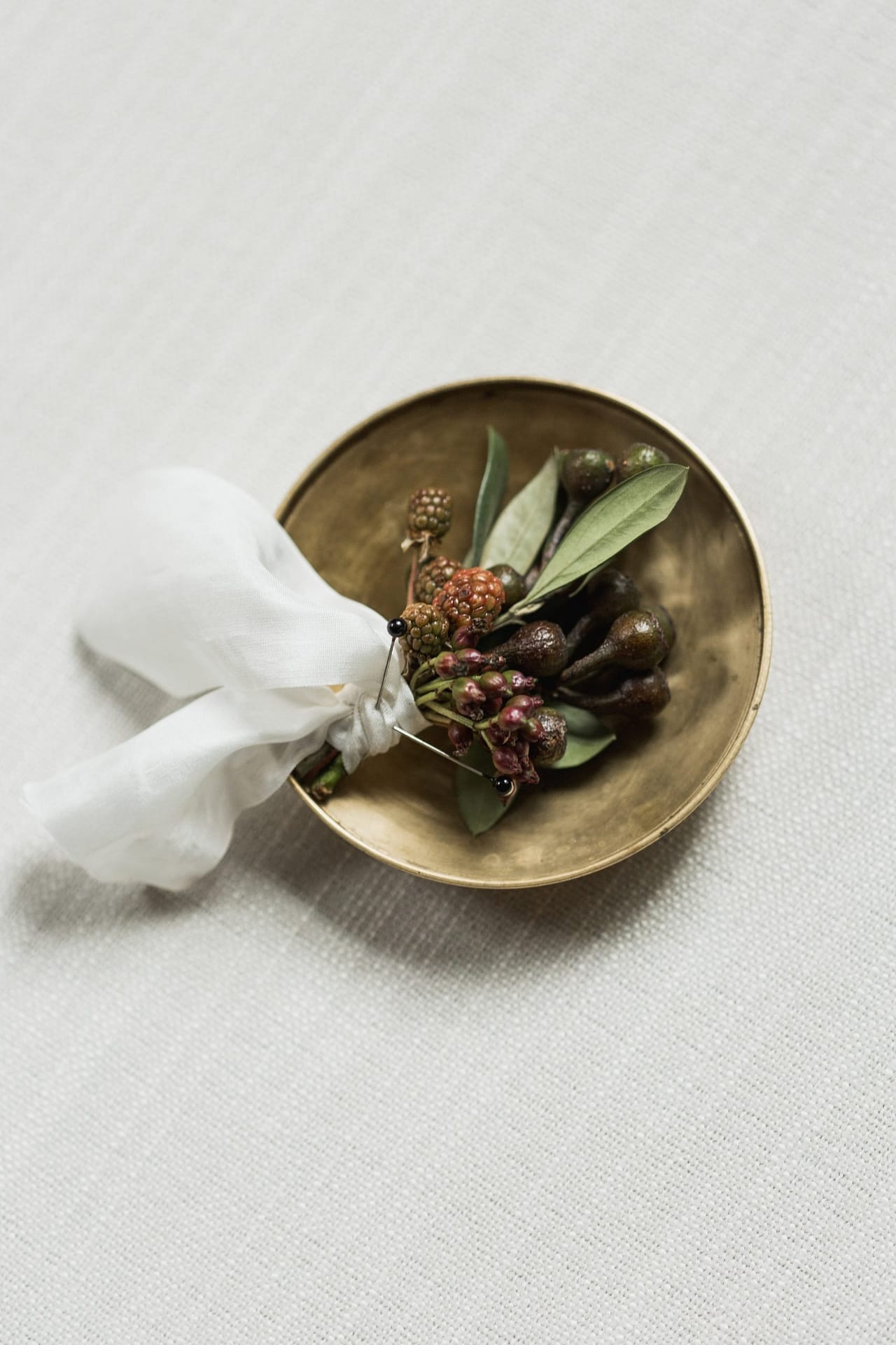 A textured boutonniere of olive foliage and berries atop a gold tray with flowing silk ribbon by Nectar and Root, Vermont wedding florist.